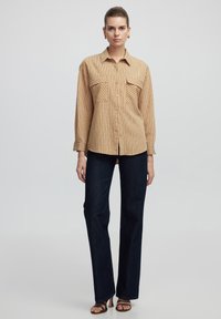 Beige striped button-up shirt with two chest pockets, paired with dark blue straight-leg jeans and brown strappy sandals. Smooth fabric texture.