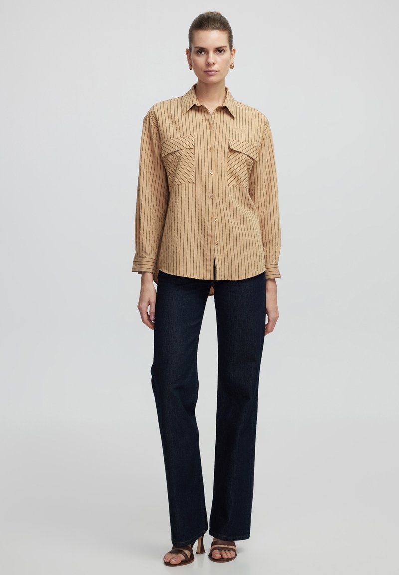 Beige striped button-up shirt with two chest pockets, paired with dark blue straight-leg jeans and brown strappy sandals. Smooth fabric texture.