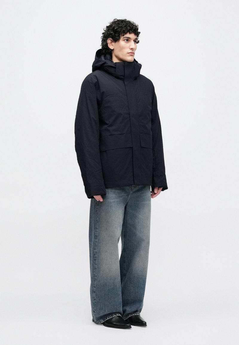 Navy outdoor jacket with a high collar and hood, featuring a zip front, two large front pockets, and long sleeves. Worn with loose-fitting denim.