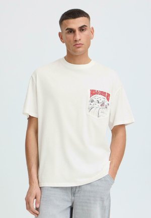 Mlad moški s kratkimi lasmi, ki nosi belo preveliko majico s rdečim in črnim grafičnim motivom na levem prsnem žepu, svetlo sive kavbojke.