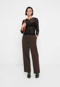 Μαύρη κορυφή με μακριά μανίκια από δαντέλα και εφαρμοστή σιλουέτα, συνδυασμένη με ψηλόμεσο καφέ παντελόνι και μαύρες μποτάκια αστραγάλου. Απλή καφέ τσάντα χειρός.