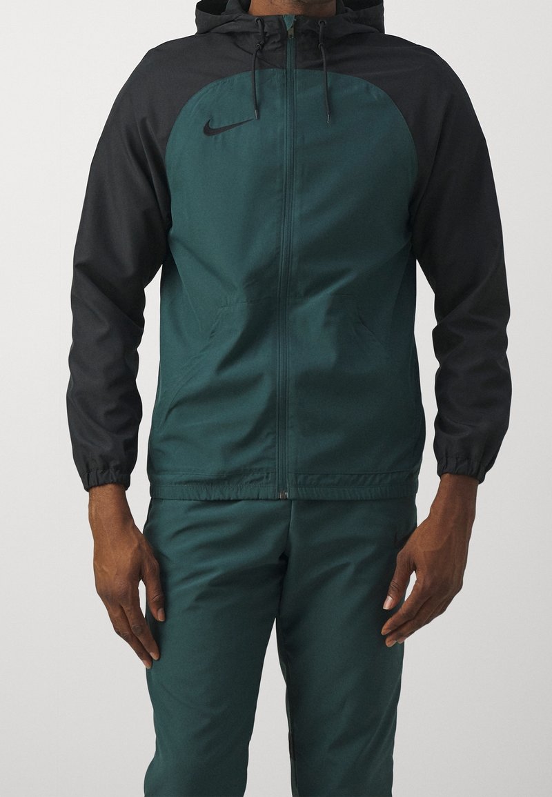 Homme portant une veste à capuche zippée avec des manches noires et un corps bleu sarcelle foncé ainsi qu’un pantalon assorti bleu sarcelle foncé.