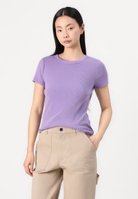 T-shirt a maniche corte viola a coste con scollatura rotonda, indossato con pantaloni beige dotati di tasche frontali e una vestibilità comoda.