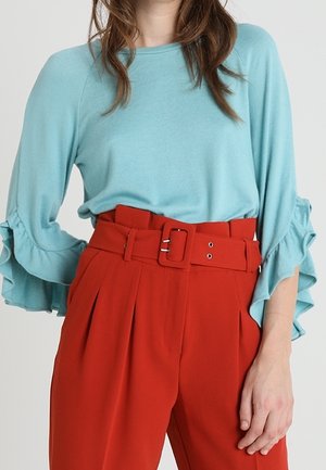 Kvinde iført lyseblå langærmet top med flæseærmer og røde bukser med høj talje, brede bælte og spænde.