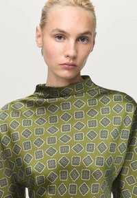 Top verde estampado con un cuello alto. Presenta un diseño de diamante repetido con acentos en azul y blanco sobre una tela suave.