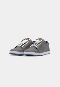 Grijze canvas sneakers hebben witte veters en een blauw-rode accent op de tong. De zool is wit met blauwe details.