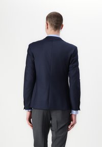 Granatowa marynarka o dopasowanym kroju, z klapami w kształcie wcięcia i jednym rozcięciem. Szare spodnie. Widać mankiety jasnoniebieskiej koszuli pod rękawami.