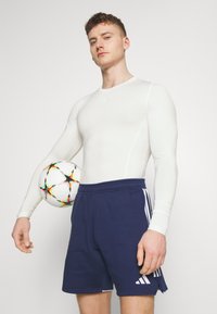 Witte lange mouwen sportshirt in combinatie met marineblauwe shorts met drie witte strepen. De speler houdt een veelkleurige voetbal tegen zijn zij.