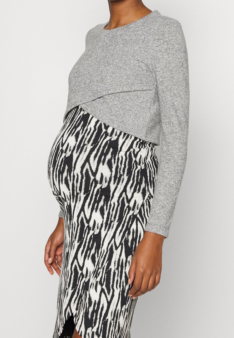Femme enceinte portant un haut gris à manches longues et une jupe ajustée à motif noir et blanc sur un fond uni.