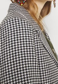 Manteau à motif pied-de-poule en noir et blanc avec une finition texturée, comprenant un col proéminent et un détail de bouton vert.