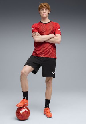 Rood gestructureerd sportshirt met zwarte accenten, zwarte korte broek, oranje voetbal schoenen en een oranje patroonvoetbal eronder geplaatst.