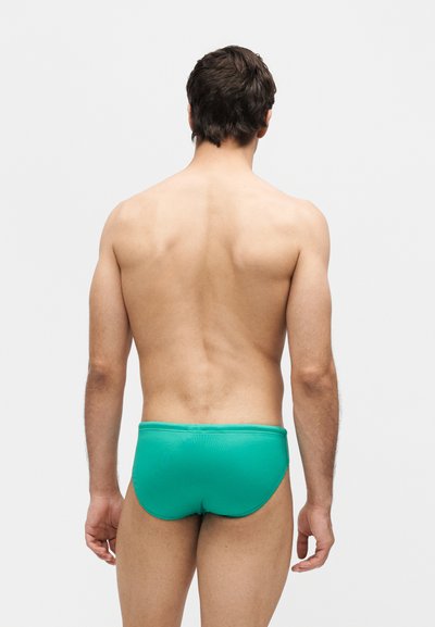 Άνδρας που στέκεται με την πλάτη, φοράει τιρκουάζ μαγιό μπροστά σε απλό ανοιχτό γκρι φόντο.