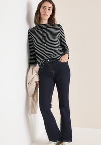 Gestreepte zwart-witte hoodie met trekkoorden, gecombineerd met donkerblauwe flared jeans met contrasterende stiksels, en een beige jack over één arm gedrapeerd.