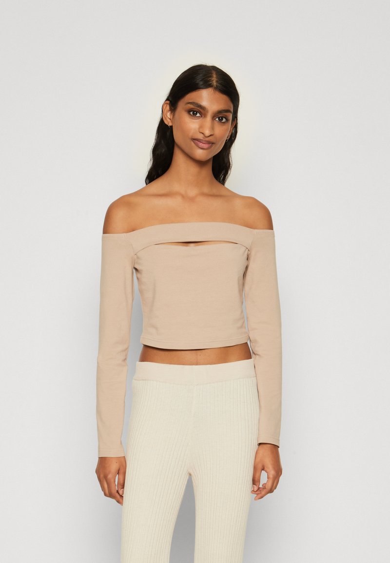 Beige avtagbar skulderkort topp med lange ermer og en frontutskjæring, laget av mykt, stretchy stoff. Kombinert med kremfargede ribbede bukser.