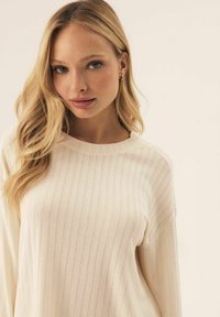 Light beige ribbed long-sleeve top with a rounded neckline and soft texture, featuring subtle vertical stripes throughout the fabric.