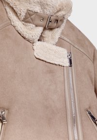 Beige suède jas met een fluffy crèmekleurige kraag, ritszakken en een leren sluiting met gesp. Zacht materiaal en moderne ontwerpelementen.