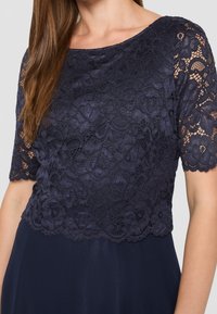 Mörkblå klänning med en spetsöverdel som har blommönster, korta ärmar och en mjuk, flytande kjol. Spetsen visar upp intrikata detaljer.