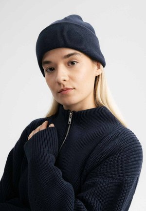 Navy knitted beanie with a ribbed texture. Worn with a matching zip-up sweater featuring a textured pattern and high collar.