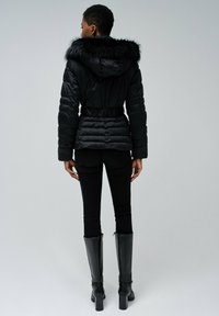 Chaqueta acolchada negra con capucha de pelo sintético, cintura ceñida y textura acolchada; combinada con jeans ajustados negros y botas altas negras.