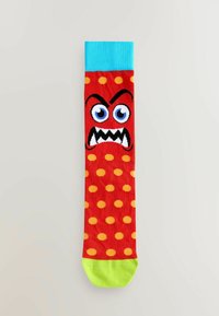 Calcetín rojo con lunares amarillos y un diseño de cara de monstruo, que presenta un puño azul y un dedo del pie verde lima, hecho de un material suave y elástico.