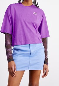 Fialové zkrácené tričko s logem Puma, vrstvené přes dlouhý rukáv s vzorem, spárované se světle modrou džínsovou minisukní.