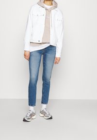 Persona vestida con una chaqueta de denim blanca, sudadera beige, jeans azules, calcetines blancos y zapatillas deRunning plateadas, de pie frente a un fondo sencillo.