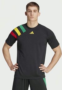 Czarna koszulka sportowa z wielokolorowym, paskowanym wzorem w odcieniach czerwieni, żółci i zieleni na lewym ramieniu, z żółtym logo na klatce piersiowej.