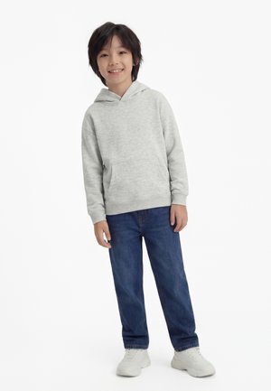 Poiss seisab, seljas helehall kapuutsiga pusa, sinised teksad ja valged tennised, naeratab tavalise valge tausta ees.