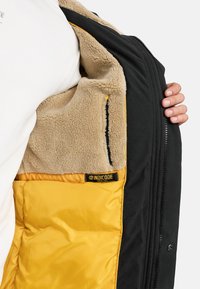 Chaqueta exterior negra con forro interior amarillo y beige. Cuenta con un interior de forro polar suave y texturizado, y una etiqueta de marca visible en el interior.