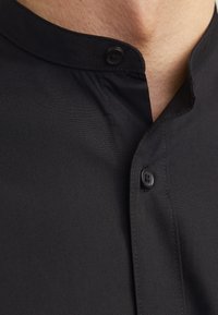 Svart skjorta med en prydlig, hög krage. Har knäppning med knappar, slät yta och en enkel design utan synliga mönster eller detaljer.