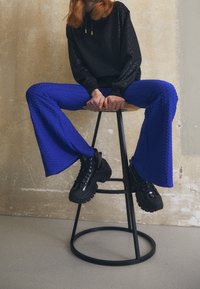 Sudadera negra texturizada con capucha, combinada con pantalones de campana vibrantes en azul que presentan un patrónhexagonal. Zapatillas negras gruesas completan el look.