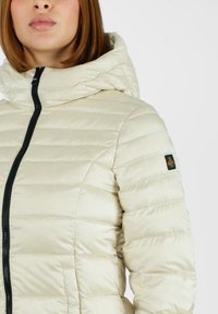 Giacca puffer beige chiaro con una texture lucida, cuciture orizzontali, una cerniera nera e un cappuccio. Presenta un logo sulla spalla sinistra.