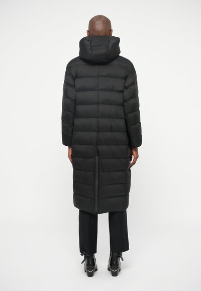 Dolga črna puhasta jakna s širokimi horizontalnimi quiltanimi odseki, kapuco in razporkom zadaj. Obuta je v črne hlače in škornje z debelim peto.