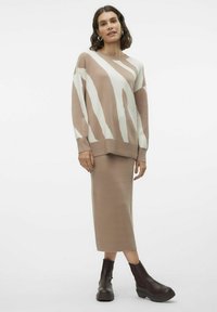 Sweter dzianinowy w beżowym kolorze z białymi paskami, zestawiony z beżową spódnicą midi. Posiada ściągacze przy mankietach i luźny krój, noszony z ciemnobrązowymi botkami za kostkę.