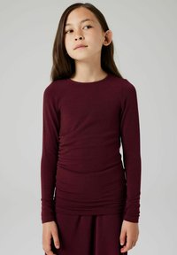 Top lungo bordeaux a maniche lunghe realizzato in tessuto morbido, con un design aderente e dettagli arricciati lungo i lati e scollatura rotonda.
