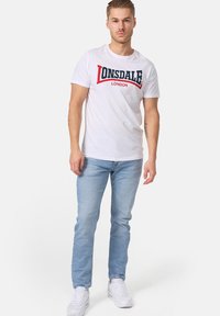 Bijela majica kratkih rukava s crveno-plavim grafičkim logotipom "LONSDALE LONDON", uparena s svijetloplavim uskim trapericama i bijelim tenisicama.