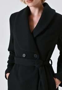 Donna che indossa un cappotto doppiopetto nero con ampi rever e una cintura legata, con le mani in tasca, su uno sfondo chiaro e uniforme.