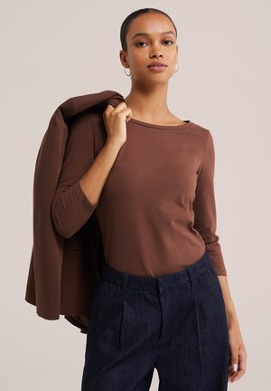 Donna che indossa una maglia marrone a maniche lunghe e pantaloni blu scuro, tenendo una giacca marrone abbinata sulla spalla contro uno sfondo semplice.