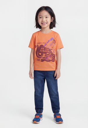 Χαμογελαστό παιδί με κοντά μαύρα μαλλιά, που φοράει πορτοκαλί μπλουζάκι με τύπωμα, μπλε τζιν με ελαστικές μανσέτες και μπλε-πορτοκαλιά αθλητικά παπούτσια, όρθιο.