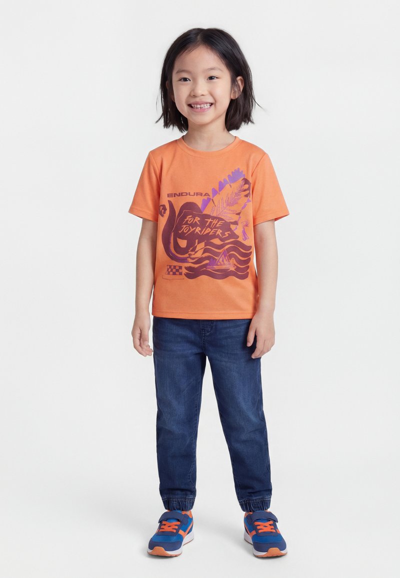 Χαμογελαστό παιδί με κοντά μαύρα μαλλιά, που φοράει πορτοκαλί μπλουζάκι με τύπωμα, μπλε τζιν με ελαστικές μανσέτες και μπλε-πορτοκαλιά αθλητικά παπούτσια, όρθιο.