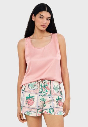 Mujer con cabello largo y oscuro, vestida con un top sin mangas de satén rosa y pantalones cortos estampados con flores, sobre un fondo liso.