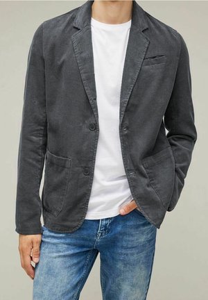 Uomo che indossa un blazer in velluto a coste grigio scuro sopra una t-shirt bianca e jeans blu, con una mano in tasca e sfondo neutro.