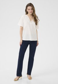 T-shirt en mélange de lin blanc avec des manches courtes et un col en dentelle ondulé, associé à un jean droit en denim foncé et des sandales à talons jaunes.