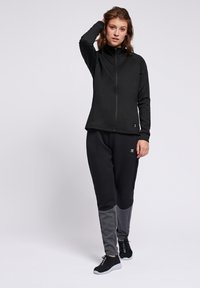 Giacca nera con zip e colletto alto, abbinata a pantaloni tapered neri e grigi. Realizzata in tessuto morbido, presenta un dettaglio del logo e pannelli in contrasto.