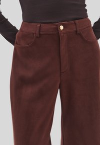 Close-up van hoge taille bruine suède broek met voorkruis zakken, riemlussen en een gouden knoopsluiting, gedragen met een donker geribbeld topje.