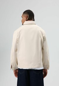 Beige gestructureerd jasje met een spreadkraag, geruite manchetten en een detail bij het rugstuk, gedragen over een wit t-shirt en donker denim.