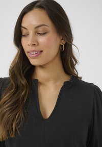 Black blouse with a V-neckline and puff sleeves, made of a textured fabric. Gold hoop earrings accentuate the look.