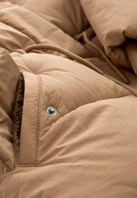 Gros plan sur le tissu d'une veste matelassée de couleur fauve, montrant un bouton-pression en métal et un cordon de serrage marron.