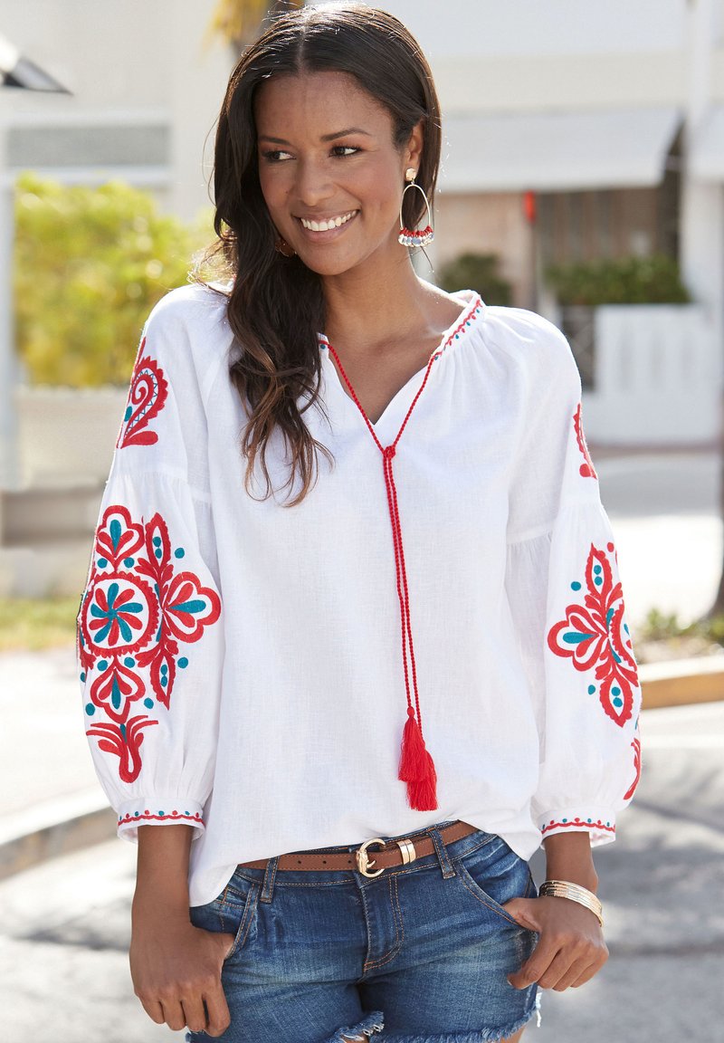 Witte geborduurde blouse met rode en turquoise bloemmotieven, oversized mouwen en een franjerugdetail, gecombineerd met denim shorts.
