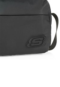 Bolso mensajero negro con un acabado de tela suave, que presenta un logotipo texturizado y una sola correa ajustable para llevar.